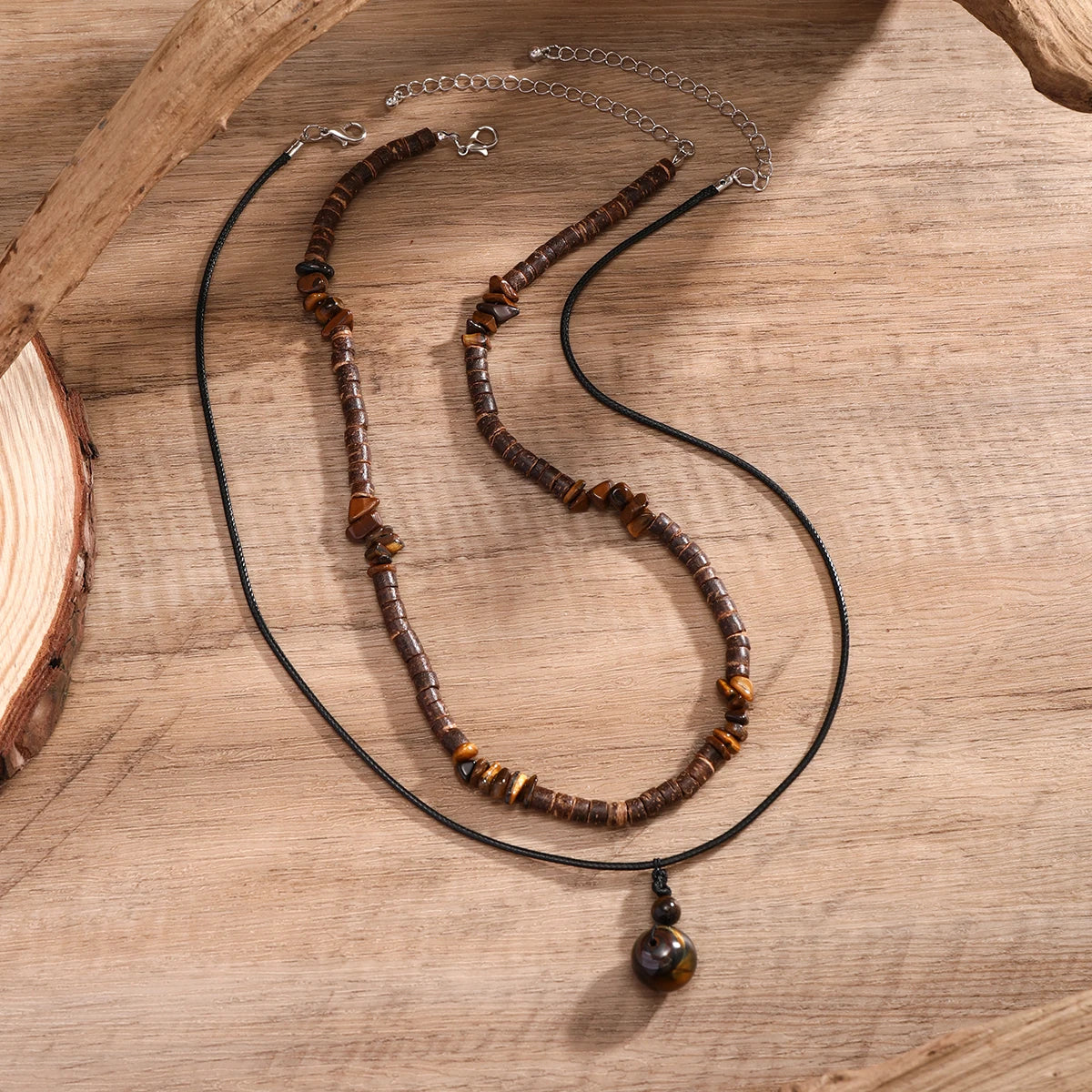 Collier perle bois homme présenté avec son second collier à pendentif, fermoirs et chaînettes visibles sur une planche en bois.
