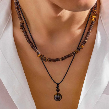 Collier perle bois homme et pendentif brun sur cordon noir, portés avec une chemise blanche ouverte, vus de trois quarts.