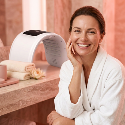 Femme souriante dans un spa lumineux aux tons rouge rosé, avec une lampe LED pour visage en arrière-plan.